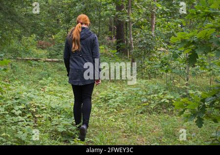 Une fille à tête rouge dans une veste noire et des leggings est marcher le long d'un chemin sur l'herbe verte dans un défrichement des forêts parmi les grands arbres avec des feuilles juteuses Banque D'Images