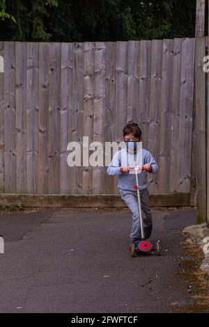 Prise de vue en longueur d'un garçon à tête rouge portant un masque facial, qui monte sur un scooter rouge sur un chemin de goudron entouré de clôtures en bois Banque D'Images