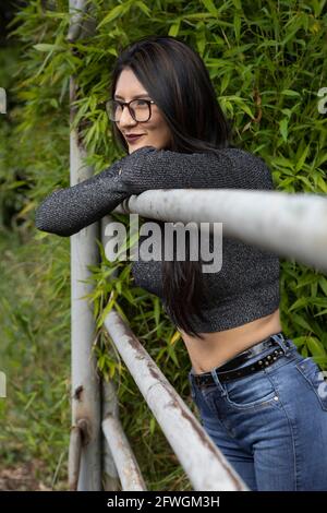 jeune femme latine avec des cheveux noirs droits portant des lunettes, des jeans et un buso décontracté gris, debout près d'une balustrade dans le champ avec des plantes, jour d'été Banque D'Images