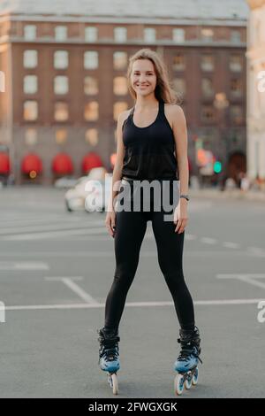 Le modèle jeune femme en vêtements de sport fait du sport sur des lames et aime les loisirs les activités se posent à un endroit urbain contre un fond flou se tient dans longueur totale Banque D'Images