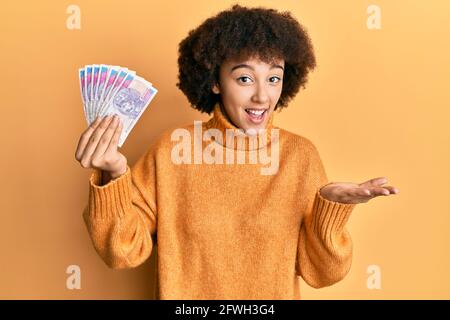 Jeune fille hispanique détenant 20 billets de banque polonais en zloty célébrant la réussite sourire heureux et expression du gagnant avec main levée Banque D'Images