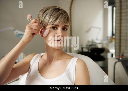 Belle femme blonde massant son visage avec un rouleau de jade assis sur une table de massage dans un cabinet de cosmétologie. Concept de soins du corps et de la peau Banque D'Images