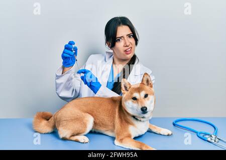 Belle femme vétérinaire hispanique mettant le vaccin à chien chiot expression sans indice et confuse. Doute concept. Banque D'Images