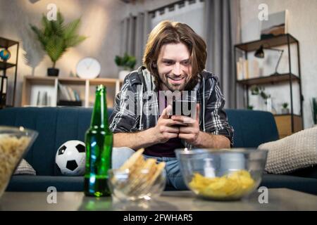 Un beau homme heureux utilisant un smartphone moderne pour regarder en direct le championnat européen de football. Un gars enthousiaste qui propose de la bière et des en-cas pour soutenir l'équipe de football préférée. Banque D'Images