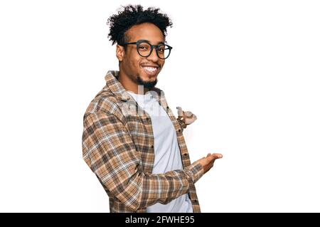 Jeune homme afro-américain avec barbe portant des vêtements décontractés et verres invitant à entrer dans le sourire naturel avec la main ouverte Banque D'Images