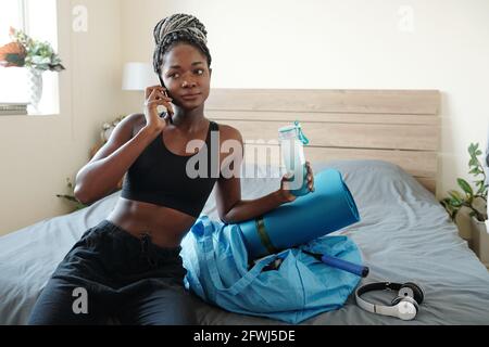 Mettre en forme une jeune femme assise sur le lit, de l'eau potable, d'emballage de sac de gym et d'appel à son entraîneur Banque D'Images