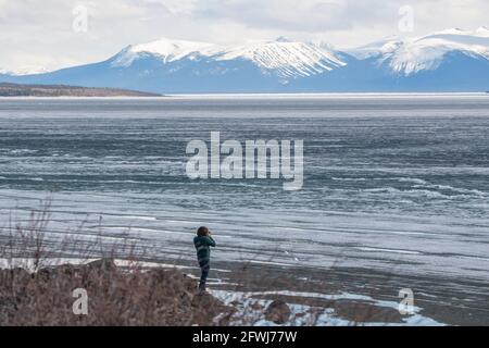 Homme debout sur la rive du lac avec de l'eau gelée partiellement dégelée avec des montagnes enneigées au loin l'après-midi nuageux. Banque D'Images