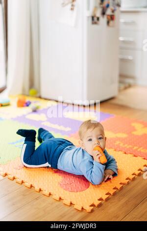 Petit garçon en costume bleu avec une carotte dans ses mains se trouve sur son ventre sur une couleur tapis sur le sol contre le fond du réfrigérateur Banque D'Images