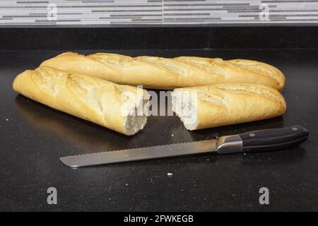 Deux pains de pain sur un banc de granit noir dans une cuisine. Une des barres est coupée et à côté est le couteau avec lequel elle a été coupée. Vous pouvez voir un allumé Banque D'Images