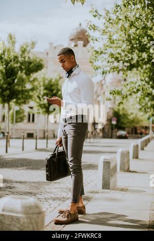 Beau jeune homme d'affaires afro-américain utilisant un téléphone portable pendant attendre un taxi dans une rue Banque D'Images
