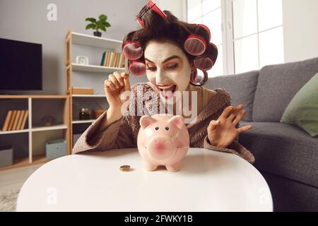 Bonne jeune femme en beauté masque facial et cheveux curlers mettre des pièces de monnaie dans une banque de porc Banque D'Images