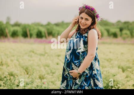 Gros plan d'une jeune femme enceinte souriante avec des fleurs sauvages à la tête en appréciant la maternité debout sur le terrain et regardant appareil photo Banque D'Images