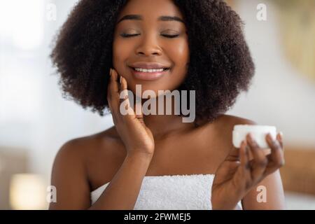 Jeune Afro-américaine appliquant de la crème hydratante sur son visage, souriant avec les yeux fermés, à l'intérieur Banque D'Images