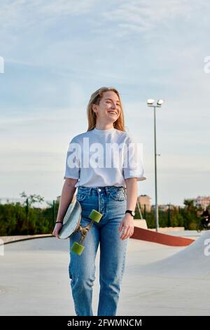 Jeune femme blonde souriant et s'amusant tenant un skateboard Banque D'Images