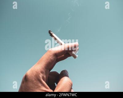 Cigarette dans la main de l'homme avec fumée sur fond bleu ciel. Tenir la cigarette à la main. Banque D'Images