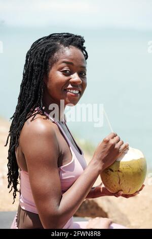 Portrait d'une jolie jeune femme qui boit un cocktail de noix de coco après l'entraînement sur la plage Banque D'Images