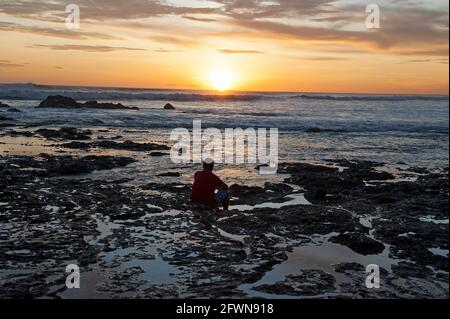 Un homme qui profite d'un coucher de soleil haut en couleur depuis un rocher à l'intérieur Des bassins d'eau à une plage au Costa Rica Banque D'Images