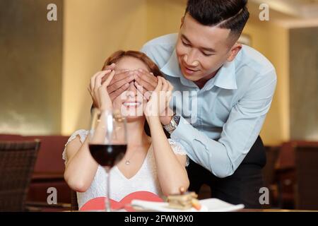 Beau jeune homme couvrant les yeux de la petite amie après l'avoir apporté délicieux dessert comme surprise Banque D'Images