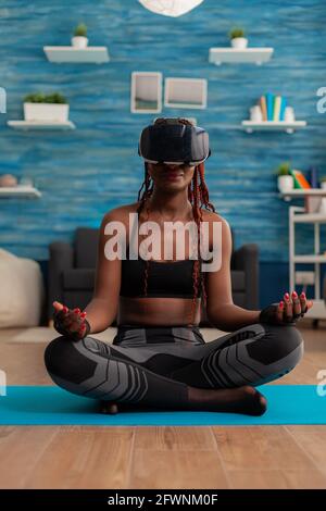 Calme tranquille femme noire portant des lunettes de réalité vrituelle assis sur le tapis de yoga, passant du temps sur la spiritualité saine mode de vie. Pratiquer la méditation dans la pose de lotus dans le salon à la maison. Banque D'Images