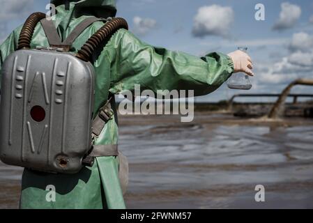 Un écologiste masculin dans un costume de protection vert et un masque à gaz prend un échantillon d'eau dans un lac pollué pour la recherche. Production de déchets. Scientifique Banque D'Images