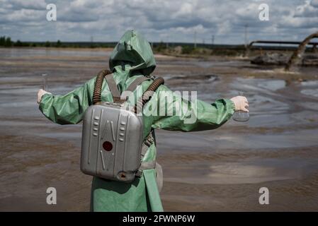 Un écologiste masculin dans un costume de protection vert et un masque à gaz prend un échantillon d'eau dans un lac pollué. Déchets de production. Un scientifique fait un Banque D'Images