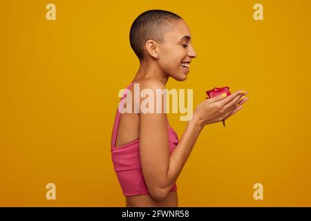 Vue latérale d'une femme rasée tenant une fleur et souriant. Femelle androgyne en casual avec une rose à la main sur fond jaune. Banque D'Images