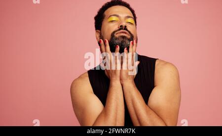 Genderqueer mâle avec maquillage d'ombre à paupières jaune debout sur fond rose. Homme fluide de genre en t-shirt noir avec ses yeux fermés. Banque D'Images