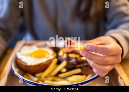 Une fille tient les frites dans le ketchup dans sa main sur un fond flou avec un hamburger. Banque D'Images