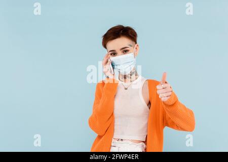 jeune femme tatouée dans un gilet orange et un masque médical parlant sur le smartphone tout en montrant le pouce isolé sur le bleu Banque D'Images