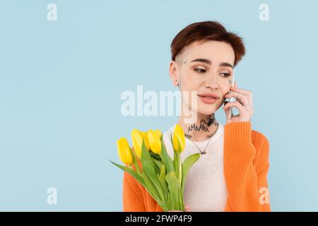 jeune femme tatouée dans un gilet orange tenant des tulipes tout en parlant sur smartphone isolé sur bleu Banque D'Images