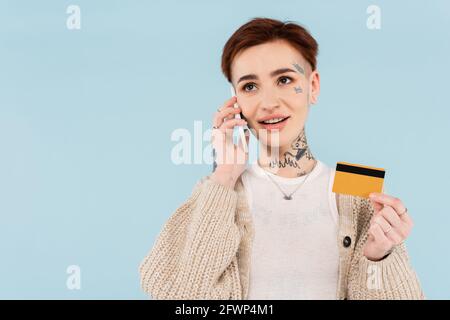 gaie et tatouée femme tenant la carte de crédit tout en faisant l'ordre sur smartphone isolé sur bleu Banque D'Images