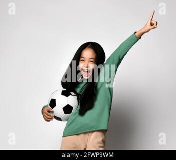 Bonne fille asiatique criant tenant le ballon de football d'une main et l'autre main vers le haut, montrant un, premier signe Banque D'Images