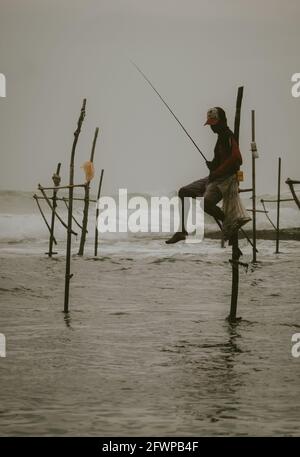 Les pêcheurs traditionnels de pilotis, qui détiennent une canne à pêche et un sac pour la collecte de poissons, assis sur une croix de bâton au-dessus de la surface de l'eau de mer patiemment waïtine Banque D'Images