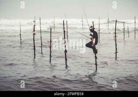 Mirissa, Sri Lanka - 07 26 2020: Pêcheurs traditionnels de pilotis tenant une canne à pêche, et un sac pour la collecte de poissons, assis sur une croix de bâton au-dessus de la Banque D'Images