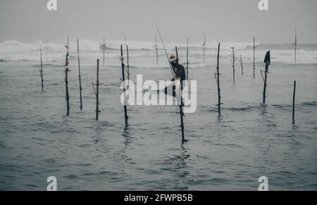 Mirissa, Sri Lanka - 07 26 2020: Pêcheurs traditionnels de pilotis tenant une canne à pêche, et un sac pour la collecte de poissons, assis sur une croix de bâton au-dessus de la Banque D'Images