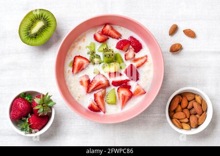 Porridge de flocons d'avoine avec des tranches de kiwi, fraises, amandes dans un bol rose, cuillère sur nappe blanche vue du dessus plat Lay Vegan concept alimentaire sain Banque D'Images