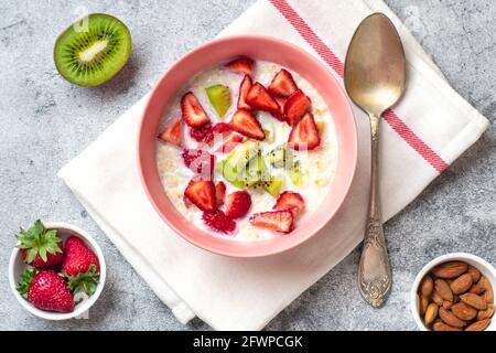 porridge de flocons d'avoine avec tranches de kiwi, fraises, amandes dans un bol rose, cuillère, serviette avec rayures rouges sur fond de béton Banque D'Images