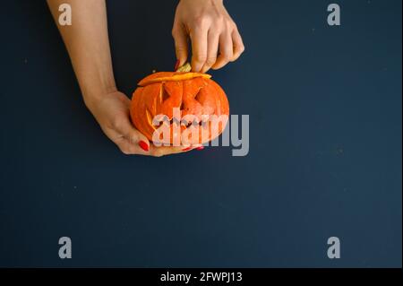 La femme tient une petite citrouille avec une grimace maléfique sur fond noir. Halloween concept, plat, vue de dessus. Le processus de fabrication de la citrouille-lanterne Banque D'Images