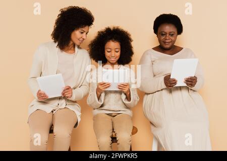 fille afro-américaine, petite-fille et grand-mère assis et regardant des comprimés sur fond beige Banque D'Images