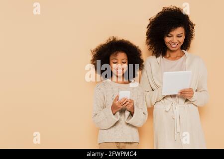 jeune fille afro-américaine souriante et mère adulte tenant un téléphone portable et tablette dans les mains isolées sur beige Banque D'Images