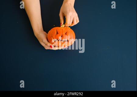 La femme tient une petite citrouille avec une grimace maléfique sur fond noir. Halloween concept, plat, vue de dessus. Le processus de fabrication de la citrouille-lanterne Banque D'Images