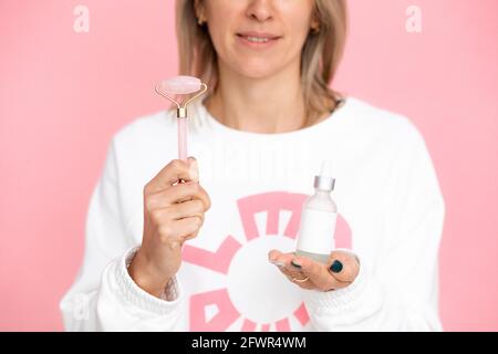 Femme blonde non reconnaissable sur fond rose tenant un rouleau de guasha et un pot en verre avec une pipette. Concept de soin du visage, alternative medi Banque D'Images