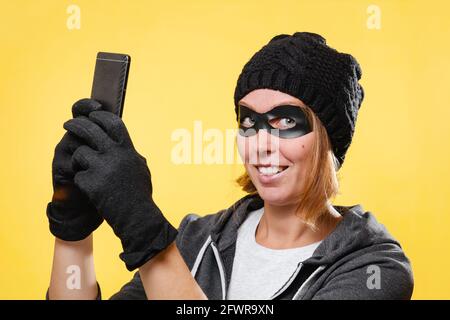 Hacker femelle dans un chapeau noir, des gants et un masque tenant un téléphone portable. Arrière-plan jaune. Concept de cybercriminalité et de piratage. Banque D'Images