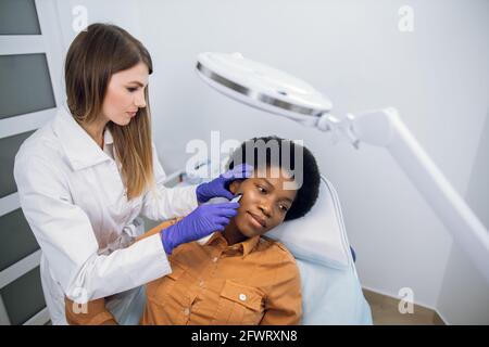 Vue de haut angle de la belle jeune femme africaine obtenant le traitement et le nettoyage de la peau par l'épurateur ultrasonique. Une esthéticienne européenne assez confiante fait un nettoyage de peau à ultrasons Banque D'Images