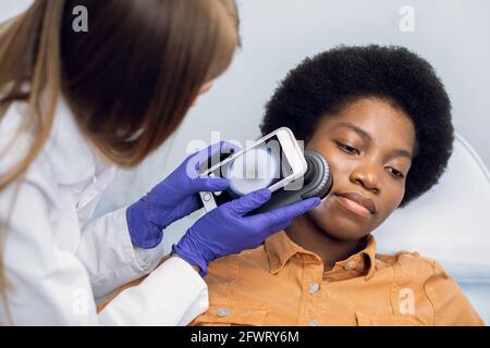 Gros plan des mains de la femme médecin tenant le dermatoscope pour l'étude des moles, des marques de naissance, des verrues, la vérification du visage de sa patiente. Belle afro-américaine couchée sur le canapé Banque D'Images