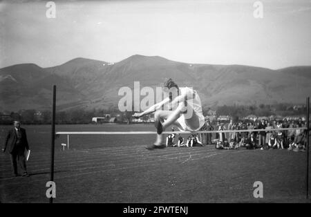 1951, image historique, sur fond de chaînes de montagnes, à l'extérieur sur un terrain d'herbe, un écolier faisant le saut en hauteur, Highlands, Écosse. Regardé par une foule de parents et d'enfants, sa technique dans sa tentative de sauter au-dessus de la barre horizontale est un saut traditionnel avec les deux pieds devant. Banque D'Images