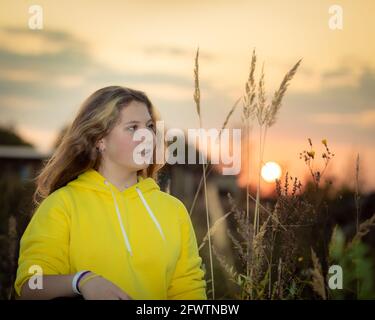 Une jeune fille avec de longs cheveux sur un pré avec de l'herbe haute dans les rayons du soleil couchant. Cheveux blonds. Veste jaune. Banque D'Images