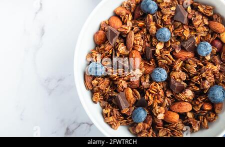 Granola au chocolat maison, muesli aux amandes, noisettes et myrtilles dans un bol sur fond de marbre blanc. Petit déjeuner sain. Vue de dessus. Pose à plat m Banque D'Images