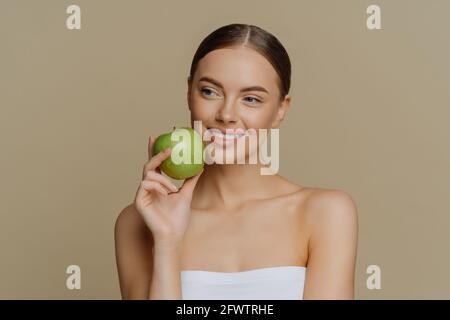 La charmante femme européenne attentionnés tient la pomme près du visage souriant doucement a des dents blanches parfaites saine peau propre enveloppée dans la douche porte-serviettes avec Banque D'Images