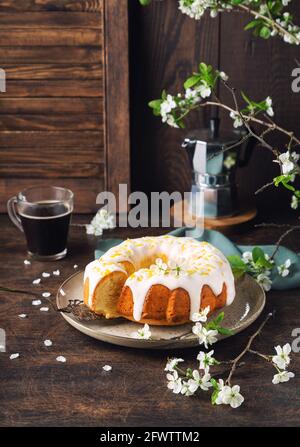 Petit gâteau au citron frais maison décoré de glaçure blanche et de zeste sur un fond rustique en bois avec des branches de prune en fleur. Banque D'Images